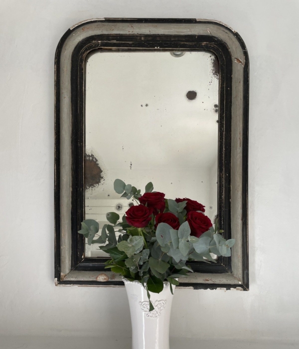 A vase of red roses and eucalyptus on a shelf in front of an antique mirror.