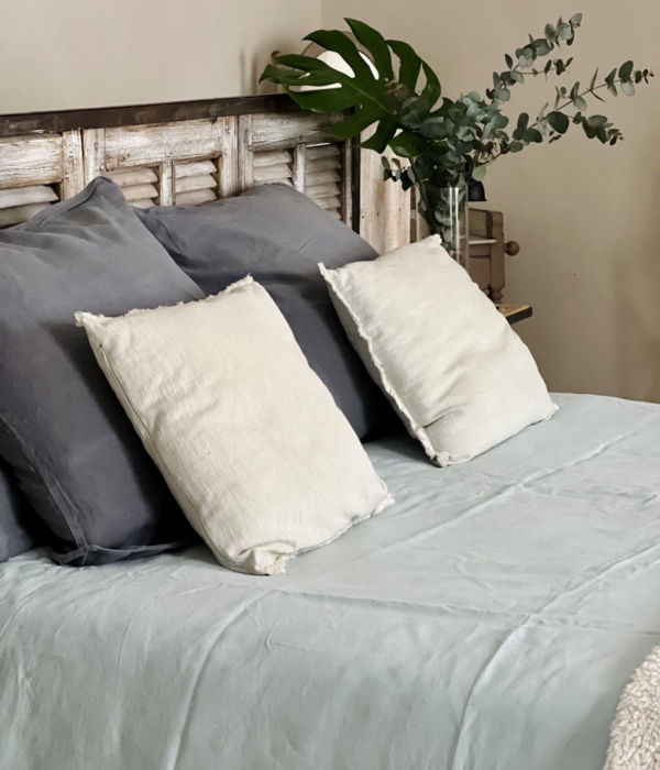 A cozy bed with grey and white pillows, pastel blue bedding and a plant.