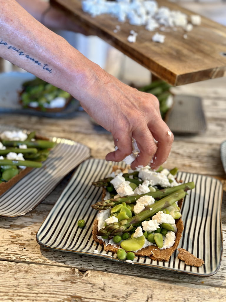 Eine Hand gibt dicke Bohnen zu einer Scheibe Spargel und Ziegenkäse – ein leckeres Gericht.