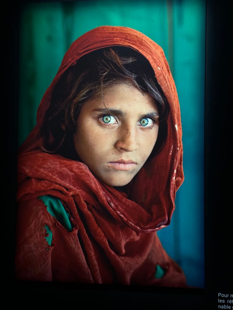 Young Afghan girl with blue eyes, wearing a red shawl against a turquoise background.