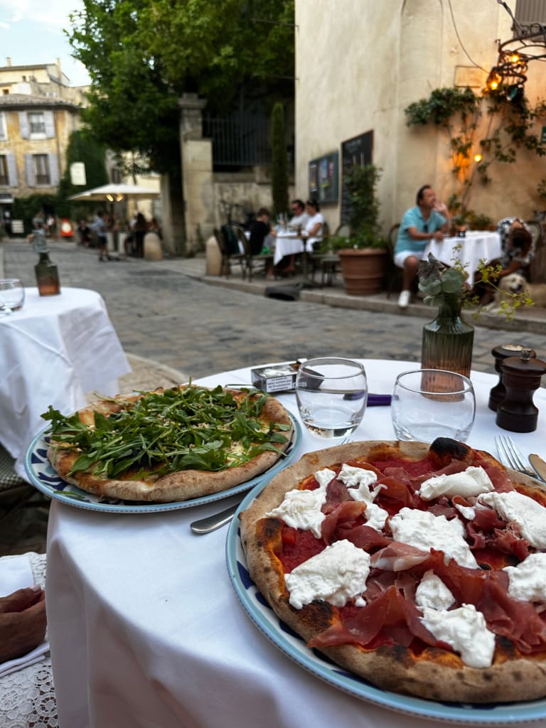 Zwei köstliche Pizzen werden auf einem Tisch auf einer Terrasse in einer malerischen Straße serviert.