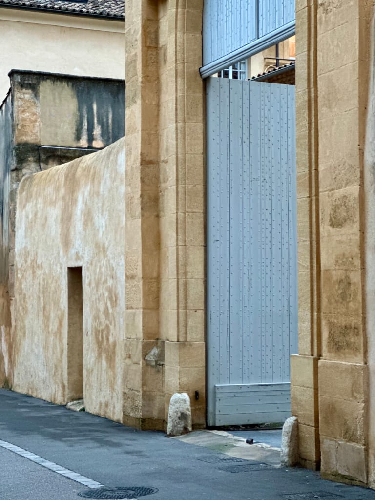 Mur de pierre beige avec une porte métallique et un bâtiment ancien.