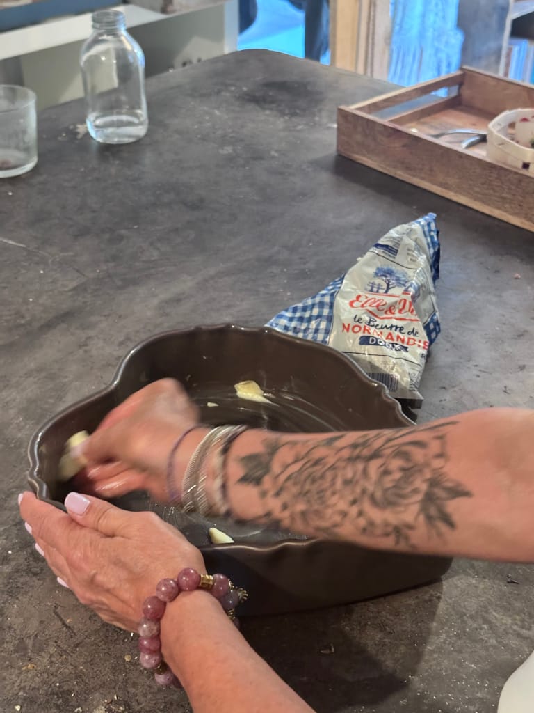 Une personne tatouée beurre un plat à tarte brun sur une table sombre.