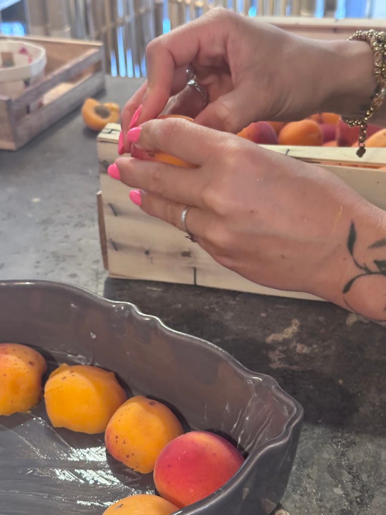 Une personne aux ongles roses manipule des abricots dans une caisse en bois et un plat gris.