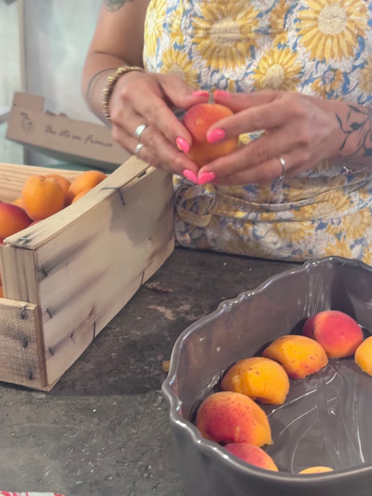 Une personne avec des ongles roses manipule des abricots entre un panier en bois et un plat en verre.