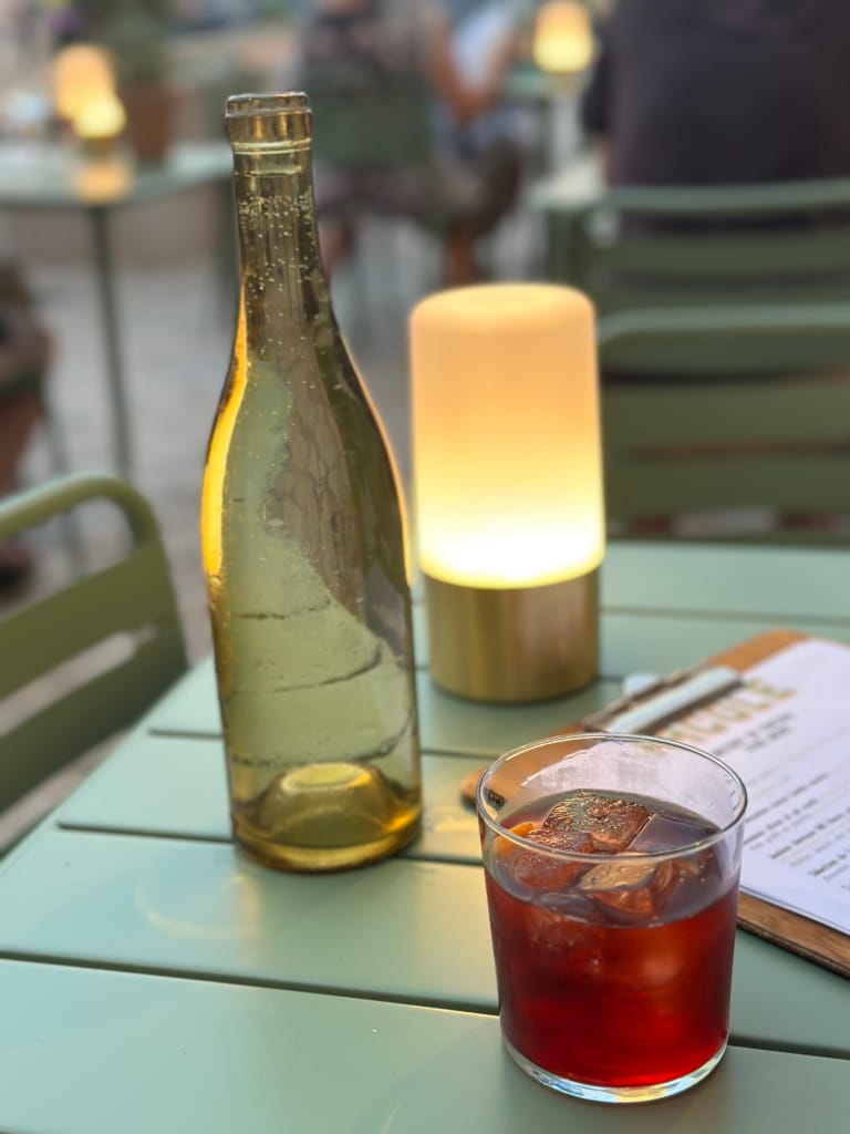 Eine Flasche Weißwein und ein roter Cocktail mit Eiswürfeln auf einem Terrassentisch.