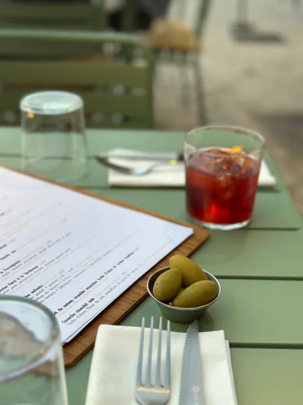 Une table de café avec un menu, un verre de cocktail rouge, un bol d'olives vertes et des couverts.
