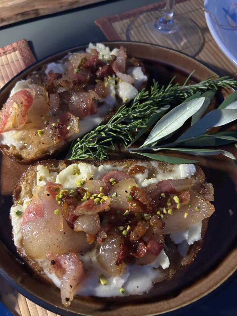Deux bruschettas garnies de fromage blanc, lardons grillés et éclats de pistaches, décorées de romarin et de sauge.
