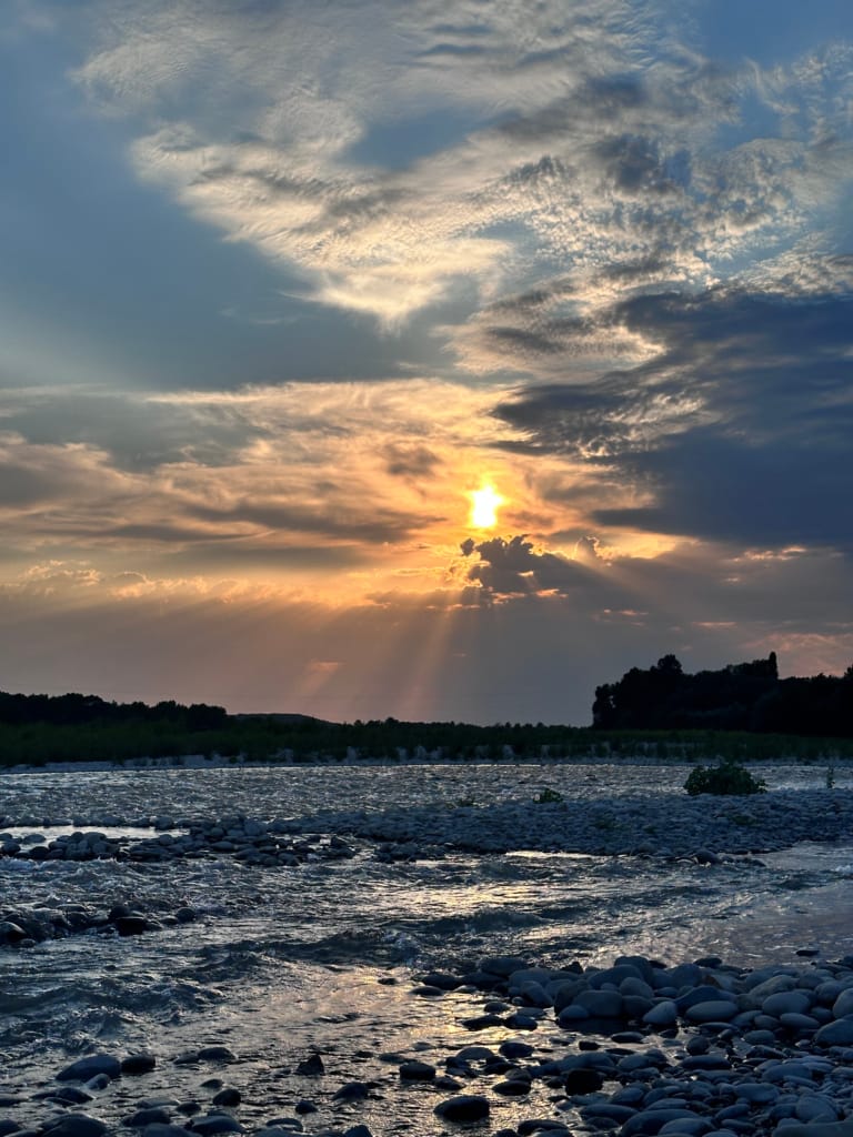 Coucher de soleil spectaculaire avec des rayons lumineux perçant les nuages au-dessus d'une rivière rocailleuse.