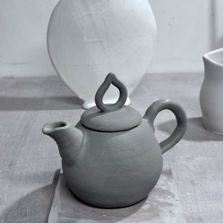 Une théière en terre cuite brute posée sur un placage, avec une jarre blanche en arrière-plan.