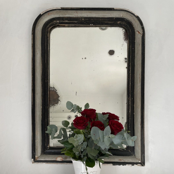 A vase of red roses and eucalyptus on a shelf in front of an antique mirror.