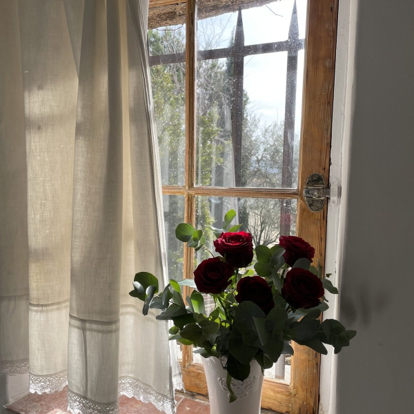 A vase of red roses in front of an old, sunny window and a light curtain.