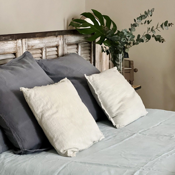 A cozy bed with grey and white pillows, pastel blue bedding and a plant.
